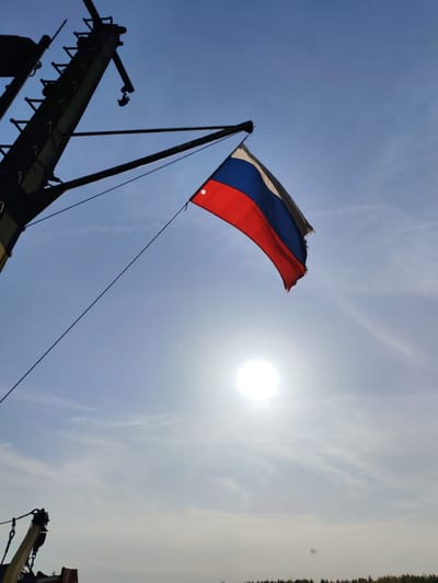 A tattered Russian flag hangs from a metal arm extending from a dark, possibly metallic, structure, potentially part of a ship or industrial equipment. The flag's colors (white, blue, red) are clearly visible. The background is a bright, hazy sky with the sun shining directly behind the flag. The scene appears to be outdoors. A thin cable runs diagonally from the structure to the area near the sun. The bottom portion of the image suggests the edge of some equipment and trees in the distance, perhaps denoting a coastal area. No people are visible in the frame. The location is specified as Shelomovo, Russia.