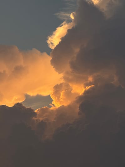 This is a vertical, close-up, low-angle shot of dramatic storm clouds at sunset over Putten, Netherlands. The clouds are dark grey and ominous in the foreground and middle ground, with streaks of bright orange and yellow light illuminating their edges and the sky behind them. The light suggests the sun is low on the horizon, likely setting, and is peeking through the dense cloud cover. The sky above the clouds is a muted blue. There are no people, animals, or structures visible. The overall impression is one of natural beauty and the powerful forces of weather.