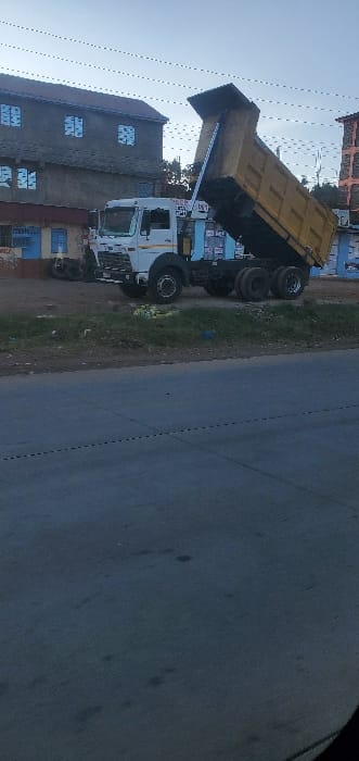 The image displays an urban street scene in Kikuyu Township ward, Kenya, featuring a white-cabbed dump truck with an elevated yellow dump bed. The truck is positioned on a dirt and grass verge adjacent to an asphalt road that occupies the foreground. The dump bed is hydraulically raised to a steep angle, extending upwards and to the right, implying a material discharge operation.

In the background, a two-story building with grey walls and a reddish-brown roof is visible on the left, featuring multiple windows with light blue frames or covers. A portion of another multi-story structure, appearing reddish-orange, is present on the far right. The grassy verge exhibits scattered light-colored debris. Overhead, several dark power or utility lines traverse the light blue-grey sky. No individuals are present in the scene, and no discernible human activity is occurring.