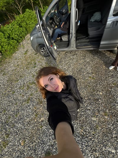 A woman is taking a selfie in a gravel lot. A grey van with open doors is behind her. The surrounding environment includes gravel, grass, and trees. The location is possibly Abzhanda, Abkhazia.