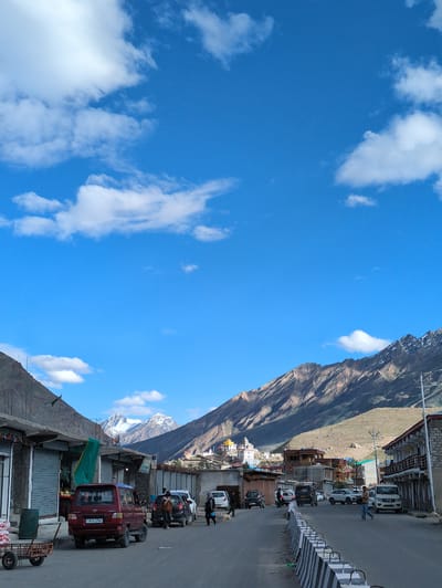 A wide-angle, full shot captures a street scene in Padum, India, beneath a vibrant blue sky dotted with white clouds. The street runs through a valley with steep, rugged mountains on either side. The mountainsides are largely barren, with some patches of scrubby vegetation. The town of Padum is visible nestled in the distance, characterized by white and yellow buildings, some with distinctive architectural features, situated on the slopes of the mountains.
In the foreground, the asphalt street is lined with shops and buildings. The shops on the left have their metal shutters down, while the buildings on the right appear to be multi-story structures with balconies. A small red SUV with license plate "JK10 2768" is parked on the left side of the street. Several other vehicles, including cars and what appears to be a white SUV, are also present on the street, suggesting moderate activity.
A few individuals are walking along the street. On the right side, a person wearing a brown jacket and blue jeans is walking away from the camera. Further down the street, another person is walking towards the camera. On the left side, near the shops, a few more figures are visible, some appearing to be near the parked red SUV. A dog is also present on the street near the cars.
To the left of the red SUV, a rustic wooden cart with wheels is parked, laden with white sacks. The street is demarcated by a concrete barrier with alternating black and white stripes, serving as a protective measure. The overall impression is a bustling, yet serene, mountain town environment under a clear, sunny sky.