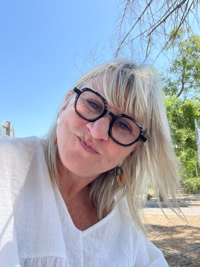 A woman with blonde hair and glasses takes a selfie outdoors. She is wearing a white top and her hair is blowing in the wind. The background shows a bright blue sky, green trees, and a building with textured walls. The sunlight suggests it is daytime, likely a sunny day. The woman has a slightly pursed lips expression, and her eyes are looking towards the camera. She is wearing a pair of black-rimmed glasses and a tortoiseshell-patterned earring. A thin gold chain necklace is visible around her neck. The setting appears to be a natural or semi-outdoor environment, possibly a garden or courtyard.
