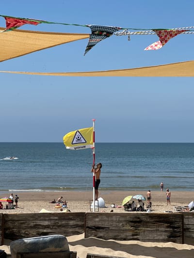 This image depicts a sunny day at a beach in Zandvoort, Netherlands. In the foreground, a wooden barrier separates the sand dunes from the main beach area. On the beach, several people are seen enjoying the day. Some are sitting under colorful umbrellas, while others are sunbathing or walking near the water. In the middle ground, a yellow flag with a warning symbol of a person in water is flying from a pole. A young person is standing on a ladder next to the flag pole, appearing to adjust it. The sea stretches out to the horizon under a clear blue sky. In the distance, a jet ski is visible on the water, and a few birds are flying in the sky. The overall scene is one of a typical summer day at a beach resort.