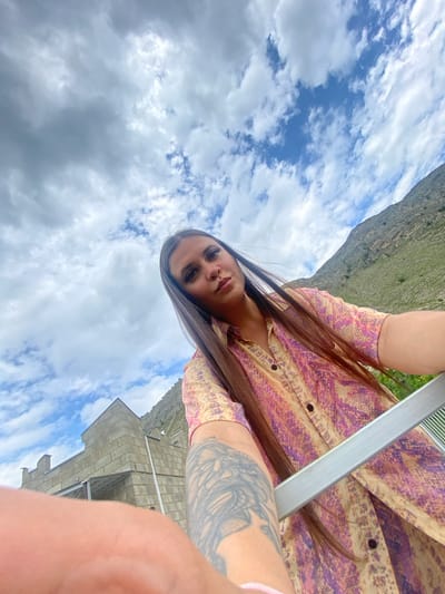 A young woman with long, dark hair and makeup is captured in a low-angle selfie. She is wearing a short-sleeved, collared shirt with a snakeskin-like pattern in shades of pink, purple, and yellow. A tattoo is visible on her left arm. She is outdoors, with a cloudy sky above. To the left, a stone building with a triangular roof and decorative turrets is visible, possibly a structure in the city of Chalda, Russia. In the background, a grassy, mountainous landscape rises up. A silver metallic object, possibly part of a railing or furniture, runs across the foreground of the image.