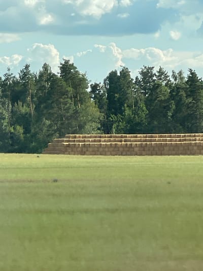 The image depicts a rural scene with a pyramid of hay bales set against a backdrop of a dense forest. The hay bales are stacked in ascending tiers, forming a triangular shape. The foreground is a grassy field, and the sky above is filled with scattered clouds. The location is identified as Pyrizhky, Ukraine, suggesting a pastoral or agricultural setting. There are no people or animals visible in the image.