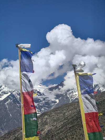The image depicts prayer flags fluttering against a backdrop of snow-capped mountains under a blue sky with white clouds. Two prayer flagpoles are visible in the foreground, with prayer flags of various colors, including blue, white, red, green, and yellow, attached to them. The flags are made of fabric and are partially torn in places. The mountains in the background are rugged and covered with snow, with a barren, rocky terrain in the mid-ground. The scene appears to be in a mountainous region, likely in the Himalayas. The presence of prayer flags suggests a Buddhist or Tibetan cultural influence. The location is identified as Kardang, India, which is a village in Lahaul and Spiti district of Himachal Pradesh. The image is a vertical, eye-level shot.