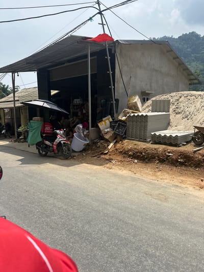 This image captures a roadside scene in Minh Sơn Commune, Vietnam, depicting a commercial area adjacent to a paved road. The main subject is a single-story concrete building with a corrugated metal roof extension, possibly serving as a shop or dwelling. To its right, a large pile of sand or dirt is visible, alongside stacks of corrugated fiber-cement roofing sheets and a wheelbarrow, suggesting construction material sales or an ongoing project.
In front of the shop, a red and white Yamaha Exciter-style scooter is parked, fitted with a red J&T Express delivery box on the back. The box prominently displays the "J&T Express" logo, along with "DỊCH VỤ PHÁT NHANH" (Express Delivery Service) and a phone number "1900 1088". Other objects near the scooter include a white plastic tub, various cardboard boxes, and a green tarp covering some goods, possibly produce like coconuts or melons. Utility poles and wires are visible in the upper left, and an orange flag hangs from the shop's roof structure.
The background features lush, tree-covered mountains under a bright, clear sky, indicating good weather, likely mid-day. The road in the foreground shows a dirt shoulder blending into the asphalt surface. The overall scene suggests a busy, functional setting common in rural or semi-urban Vietnamese areas, bustling with daily commerce and activity. The red object in the immediate foreground right is likely part of the viewer's motorcycle.