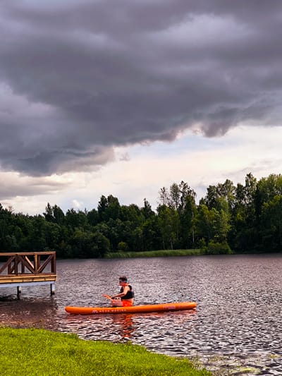 An individual wearing an orange life vest and black shorts is paddling an orange stand-up paddleboard on a body of water. The paddleboard is labeled "AQUA MARINA FUSION". The person is positioned centrally on the board, facing towards the left of the frame, with a paddle in hand. In the background, a dense line of green trees forms the shoreline. To the left, a wooden dock with a railing extends into the water. The sky is overcast with dark, dramatic clouds, suggesting an impending storm. The foreground features a strip of vibrant green grass. The overall scene depicts an outdoor recreational activity on a lake or river with moody weather conditions. This scene is consistent with the natural landscapes found around Vecumnieki, Latvia, known for its numerous lakes and forests.
