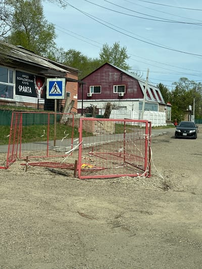 The image shows a street scene in Raychikhinsk, Russia. A red metal barrier sits in the foreground on a gravel road, secured with what appears to be white tape. Behind the barrier is a building, on which a sign that includes the words "APOO" and "SPARTA" can be seen in the lower left corner, as well as a pedestrian crossing sign. Further down the road are two more buildings, including one with a steeply pitched roof with red and white panels. In the background, a black car is parked. Overhead are power lines.