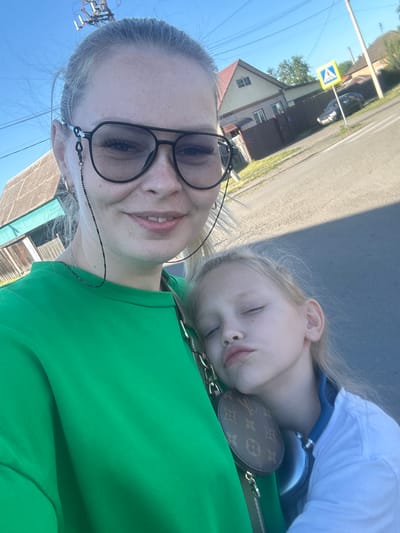 In an outdoor, daytime scene in the city of Kalinino, Russia, a blonde woman wearing large, dark-rimmed glasses and a bright green sweatshirt is taking a selfie. She is in the foreground, looking at the camera with a slight smile. Her hair is pulled back, and a dark beaded chain for her glasses hangs down. She is holding a young blonde girl who is leaning her head on the woman's shoulder, eyes closed, with a pout. The girl is wearing a white collared shirt. A small, round coin purse with a Louis Vuitton pattern is visible, hanging from the woman's side. In the background, there are houses with wooden fences, a paved road, a pedestrian crossing sign, a car parked on the side of the road, and utility poles with wires overhead. The sky is clear and blue.