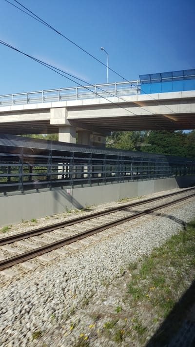 The image shows a modern overpass with a light pole and overhead electrical wires against a clear blue sky. Below the overpass is a train track with ballast and a long, grey structure with metal fencing and translucent panels. Green trees are visible behind this structure. The train tracks run diagonally across the foreground, with some patches of grass and small yellow flowers growing between the ballast. The scene appears to be in Teplička nad Váhom, Slovakia, likely near a transportation hub or a construction site given the infrastructure.