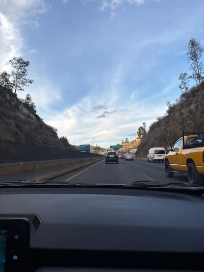 View from inside a vehicle on a highway. There is traffic ahead, with cars, vans, and a yellow truck visible. A hillside lines one side of the road, and the sky, with scattered clouds, is visible overhead. Part of the vehicle's dashboard and a mounted device are in the foreground.