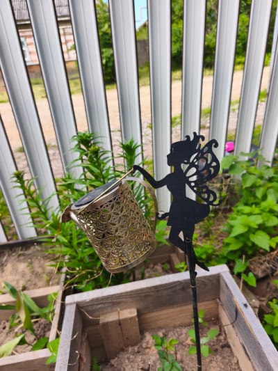 A black silhouette of a fairy with detailed wings and a crown stands on a thin metal rod. The fairy is positioned to the right of a decorative, golden watering can. The watering can is cylindrical with an intricate, perforated pattern and has a solar panel on top. It is tilted as if pouring water, with wispy, dried plant tendrils hanging from its spout. The scene is outdoors, with a white, corrugated metal fence in the background. Behind the fence, a brick building with a light blue window is partially visible, along with green foliage and trees. The foreground shows a raised garden bed made of weathered wooden planks, filled with soil and small green plants. The location is Puchezh, Russia.