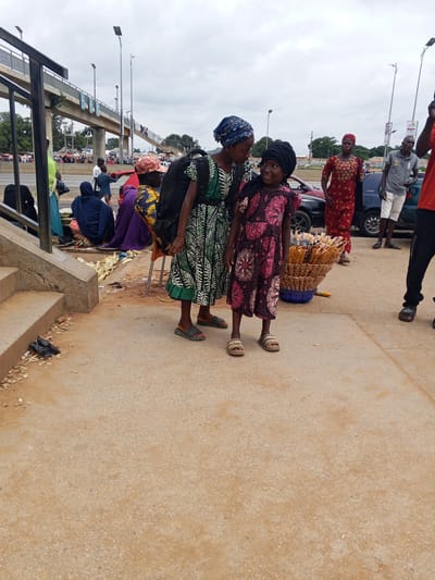 This media file captures a scene on a sidewalk in Immigration Quarters, Nigeria. The primary subjects are several individuals, mostly women and children, dressed in vibrant African attire. In the foreground, two young girls are interacting, with one older girl looking down at the younger one with a gentle expression. The younger girl is wearing a dark pink and white patterned dress and a black headscarf, while the older girl has on a green and white patterned dress and a blue headscarf.
Behind them, other people are visible. A woman in a red and yellow patterned dress stands to the right of the girls. Further back, a man in a grey polo shirt and white shorts is seen. Another man in a red shirt and black pants is also present on the right side of the frame. Several other individuals are scattered in the background, some sitting or standing.
The setting appears to be a busy roadside or market area, indicated by the presence of vehicles (a dark red car and a dark blue car are partially visible) and a basket full of snacks or goods, possibly corn on the cob, being sold by one of the women. There is a pedestrian overpass visible in the background, suggesting the location is near a major road. The ground is paved with concrete and dirt.
The weather appears to be overcast, with a cloudy sky. The time of day is not explicitly indicated but the lighting suggests it could be daytime. The overall mood seems casual, with the interaction between the girls conveying a sense of warmth and connection. Some debris and discarded items are visible on the ground. No legible text is clearly discernible in the image.