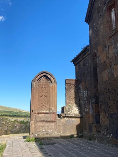The image captures an exterior view of an ancient stone structure, possibly a church or monastery, against a clear, bright blue sky. The stonework is weathered and exhibits intricate carvings, suggesting historical significance. One prominent feature is an arched stone slab with detailed relief work, including what appears to be a cross. To the right of this, another section of stone wall is visible, adorned with geometric patterns. The ground in the foreground is paved with reddish-brown bricks, forming a walkway. Beyond the stone structure, a gently sloping landscape of green fields stretches out under the daylight. The overall impression is one of antiquity and serene natural surroundings, likely in a rural or historical location, possibly Saghmosavan, Armenia. The lighting suggests it is daytime, with the sun casting distinct shadows on the stone surfaces, indicating it's not midday. There is no visible text in the image.