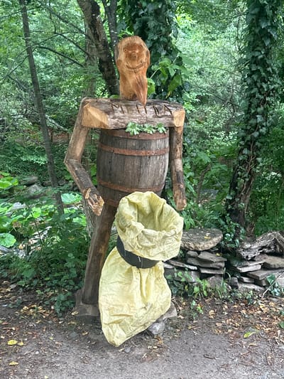 A wooden sculpture of a figure stands outdoors in a forest setting. The figure is constructed from rough-hewn wood, with a barrel serving as its torso. The head is carved from a separate piece of wood and features a smiling face with carved eyes and a wide grin. Its arms are made from branches, and it appears to be wearing a large, light-yellow bag that is cinched around its waist with a black band. The sculpture is situated on a dirt path surrounded by lush green foliage, trees, and rocks. The background shows a dense forest with ivy climbing up some of the trees. The overall impression is of a whimsical, rustic sculpture placed in a natural environment. This sculpture is located in Golyamo Belovo, Bulgaria.