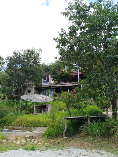 A medium shot captures an outdoor scene in Xuân Sơn, Vietnam, featuring a multilevel structure nestled amongst lush vegetation. The main focus is a large, parabolic satellite dish, partially covered by a white translucent fabric, positioned on a grassy slope in the foreground. To the right of the dish is a concrete structure that appears to be a fireplace or barbecue pit. A wooden table with a metal top sits further to the right, amidst dense green foliage. The background reveals a modern building with multiple balconies and glass windows, constructed on a terraced hillside. Several trees with green leaves are scattered throughout the landscape, and the sky above is bright, with a few scattered clouds. The ground in the immediate foreground is a mix of gravel and dirt, with scattered stones. No people are visible in the frame.