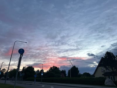 The image depicts a street scene in Laagri alevik, Estonia, during a twilight hour, with a vibrant sunset coloring the sky. The sky is filled with a multitude of clouds, ranging from dark grey to hues of pink and purple, indicating the setting sun. In the foreground, a paved road is visible with a sidewalk bordering it. Several streetlights and traffic signs are present, including a sign with a bicycle and pedestrian symbol, and another indicating a speed limit of 40 km/h. Trees and dense green hedges line the street, with a residential building visible on the right side of the frame, featuring a sloped roof and windows. A flagpole stands without a flag. The overall atmosphere is calm and serene, characteristic of the end of the day.