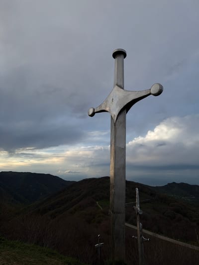 A low-angle, outdoor photograph depicts a large, metal sword monument set against a backdrop of a cloudy sky and a mountainous landscape in Zemo Nichbisi, Georgia. The sword's hilt features rounded pommels and a prominent cross-guard. Further down a metal path are two smaller metal swords. The environment consists of dark, tree-covered mountains under a sky with visible clouds and varying light conditions.
