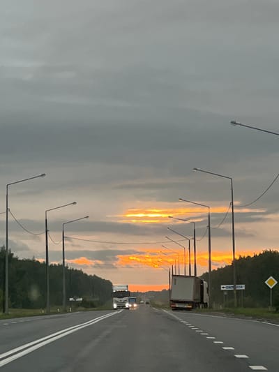 The image depicts a highway in Nadezhdinka, Russia, at dusk. The sky is a mix of gray clouds and vibrant orange sunset streaks, indicating either the end of a stormy day or an impending storm. The road, marked with white lines, curves to the right. On the left side of the road, a series of streetlights with curved arms are evenly spaced.
In the distance, several trucks are visible on the highway, their headlights on. The lead truck appears to be a white semi-trailer truck. A yellow diamond-shaped road sign is visible on the right side of the road, next to two blue directional signs. One of the blue signs has white text and an arrow pointing left. The other blue sign has white text and indicates "7 МКC".
The overall scene evokes a sense of travel and the transition from day to night. The presence of the trucks suggests ongoing transportation activity on this route. The weather appears overcast with dramatic sky colors.