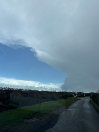 The image captures a rural roadside scene in Stratford-on-Avon, United Kingdom, under a transitioning sky. The upper left portion of the sky is clear blue with sparse white clouds, sharply demarcated from a large, heavy mass of dark grey clouds dominating the upper right. A dense, dark hedge line, appearing bare or with minimal foliage, spans the mid-ground, obscuring the distant horizon. In the foreground, a narrow, wet asphalt road curves gently to the right and slightly uphill. To the left of the road, a green grassy verge exhibits areas of mud and standing water, with several small yellow flowers visible. Further down the road, a junction is apparent on the right, marked by a red-bordered "GIVE WAY" sign, a yellow rectangular sign positioned beneath it, and an orange traffic cone. No individuals or active subjects are present in the visible scene.