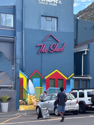 The image is of "The Shed" in Muizenberg, South Africa, an "Olympic Outlet Store," featuring painted decorations of houses. A person is standing next to a parked silver sedan, license plate "CA 976-162". Behind the sedan is a white SUV with a visible license plate that reads "KR 42". In front of the sedan is a sign that advertises "JACK BLACK ICE COLD lager". The building facade is a dark blue, with painted houses in yellow, red, and blue. There are two windows on the upper level and one on the lower level of the building. The top of a mountain can be seen in the background. There are power and cable wires running along the front of the building, along with pipes. A potted plant is on the sidewalk. Another person is standing in front of the painted wall. The building features a pink neon sign that spells out "The Shed".
