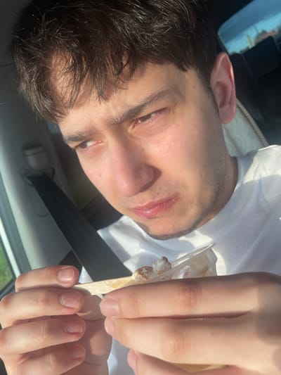 The image shows a close-up of a young person, possibly a male, inside a car. The person is looking towards the camera, with a portion of their face, including one eye, nose, and mouth, visible. They have dark hair and a light complexion. In the foreground, the person's hands are holding a small wooden stick with some white substance on it, possibly food or ice cream. One hand is touching the stick, while the other appears to be resting on the person's chin or neck area.

The setting appears to be daytime, with sunlight casting shadows, suggesting it might be a sunny day. The background shows a blurred view of the car's interior, including a seatbelt, and a reflection in a window that hints at an outdoor environment with some greenery. The overall mood seems casual, with the person possibly enjoying a snack. There is no visible text in the image. The provided location context "Гавриловка, Russia" suggests the image was taken in that region.