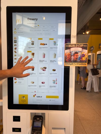 The image shows a person's hand reaching out to touch a touch-screen ordering kiosk at a McDonald's restaurant. The kiosk displays a menu of desserts, including McFlurrys, shakes, and McPops, with prices listed in Polish Zloty. A banner promoting a "FIFA World Cup Meal" with collectible cups is prominently displayed. In the background, other customers can be seen interacting with similar kiosks. The setting appears to be indoors, with bright, artificial lighting. The overall scene suggests a typical fast-food ordering experience. The visible text includes product names and prices in Polish, as well as promotional material for the World Cup. The presence of the payment terminal on the right side of the image indicates that transactions are being processed. The location is identified as Katowice, Poland.