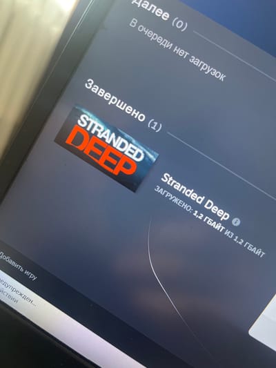 The image shows a computer screen displaying a software interface, likely a game launcher. The text is in Russian. At the top, it says "Далее (0)" which translates to "Next (0)", and below it, "В очереди нет загрузок" meaning "No downloads in queue." Further down, it says "Завершено (1)" or "Completed (1)", indicating one item has been finished.
Below "Completed (1)" is a game cover for "STRANDED DEEP." To the right of the cover, the game title "Stranded Deep" is displayed again, followed by "ЗАГРУЖЕНО: 1,2 ГБАЙТ ИЗ 1,2 ГБАЙТ," which means "Downloaded: 1.2 GB out of 1.2 GB." This signifies the game is fully downloaded.
In the lower left corner, text reads "Добавить игру" (Add game) and "Редупрежден..." which is likely "Unverified..." or "Pre-verified..." suggesting an action or status related to adding games.
The image appears to be taken at an angle, capturing a portion of the screen and its immediate surroundings, including the edge of a laptop or monitor. There are no people or clear environmental elements to suggest a specific location within the city of Sorochinsk, Russia, or any other city. The content on the screen is digital and generic to computer software.