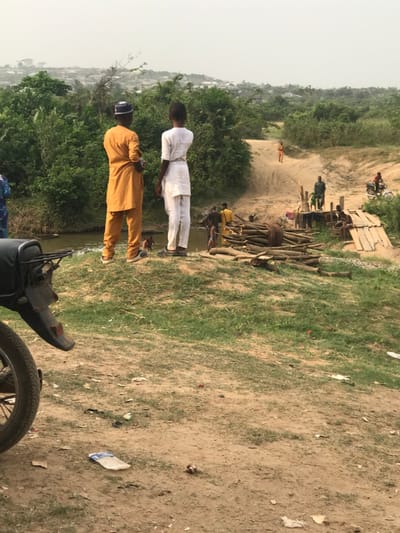 A landscape scene featuring two men in traditional attire. One wears a mustard-colored suit and cap, the other, white. They stand near a motorcycle. Behind them, a group gathers near stacked logs and a makeshift structure. Further away, other people are visible on a dirt path, with a motorcycle in the distance. The terrain consists of grass and dirt, with scattered trash. Trees and buildings are seen in the background under a cloudy sky.