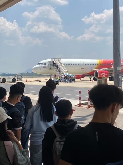 The image shows a group of people at an airport in Tambon Hua Toei, Thailand, watching passengers disembark from a Vietjet aircraft. The plane is red and white. Passengers are using a stairway connected to the front of the plane. Ground vehicles are visible near the plane. The sky is blue with scattered clouds.