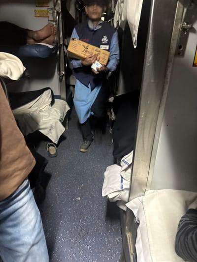 An interior scene, seemingly inside a train, depicts a cluttered space with multiple people. One individual in a blue uniform lies on a lower berth, holding a cardboard box labeled "Fresca." The lower berth also accommodates another person's feet. Unoccupied bedding and clothing are scattered around. A leg clad in denim jeans is also visible, and a metal rail suggests the proximity of other berths. The general impression is of a cramped, shared sleeping area.
