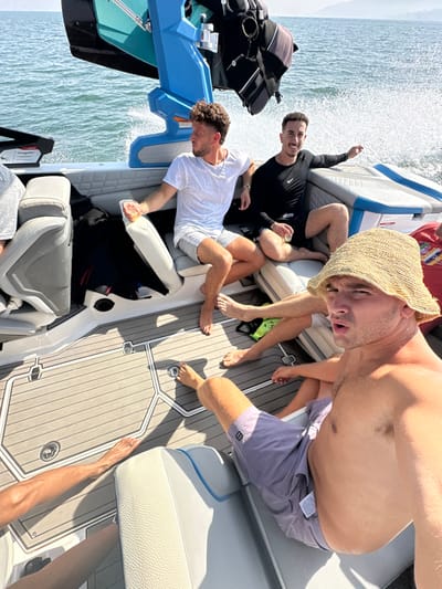 A group of young men are observed on a recreational motorboat traversing the Sea of Galilee (Lake Kinneret) near Migdal, Israel, under a clear, sunny sky. The modern boat features grey and white cushioned seating and a textured grey deck. A blue and black wakeboard tower with attached equipment is visible at the rear.
In the foreground, a shirtless male with fair skin, wearing a straw bucket hat and light purple swim shorts, looks directly towards the camera with an open-mouthed expression. To his left, another male with curly brown hair and a beard, dressed in a white t-shirt and light-colored shorts, is seated, looking towards the left side of the frame. Adjacent to him, a third male with dark hair and a beard, wearing a black long-sleeved athletic shirt with a Nike logo and black shorts, smiles towards the camera, extending his right arm and holding a small container. Bare feet and legs belonging to additional, partially visible individuals are present on the boat's deck and seats. A black item, possibly clothing, is stowed on a seat. The boat's movement generates white wake on the blue water, with distant land visible on the horizon.