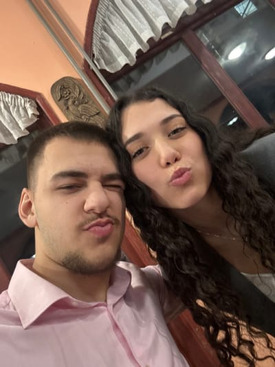 This image depicts a close-up, indoor selfie of two individuals, identified as being in Arkovna, Bulgaria. A male subject, positioned on the left, has short dark hair, a light mustache, and a chin strap beard, wearing a light pink collared shirt. He is making a facial expression with his right eye winked closed and lips pursed into a pout. To his right, a female subject with long, dark, curly hair is visible, wearing a dark outer garment over a white top. She is also looking towards the camera with her lips pursed in a pout. Both individuals occupy the majority of the frame. The background shows an interior setting with light orange walls, dark reddish-brown wooden trim, white ruffled curtains visible at the top, and a carved wooden decorative panel between and behind the subjects. Reflections of interior lighting are present in a window or glass panel on the right side of the image.