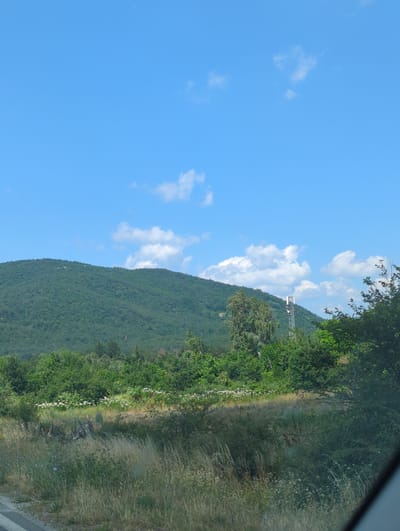 This media file depicts a landscape view from a vehicle, oriented vertically. The scene is dominated by a lush, green mountainside on the right, stretching into the distance. The mountain is covered in dense vegetation, with trees and shrubs forming a textured surface. A thin, white line of flowers or small plants runs along a path or the edge of the road on the lower part of the mountain. To the left, a vast, clear blue sky with scattered white, fluffy clouds fills the frame. A tall, lattice-like communication tower is visible on the upper part of the mountainside, silhouetted against the sky and clouds. The overall impression is of a bright, sunny day, possibly late morning or early afternoon, given the strong sunlight illuminating the scene. The location is identified as Gurkovo, Bulgaria, suggesting a rural or mountainous setting. There are no people or specific activities evident, but the perspective implies someone is traveling and capturing the scenery. The image has a somewhat reflective quality, possibly indicating it was taken through a window.