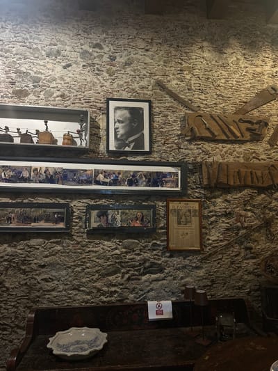 The image depicts a stone wall in an indoor setting. Various framed pictures and objects adorn the wall. There is a black and white photograph of a man's face, a newspaper clipping with the headline "Il Padrino," and several framed photographs, one appearing to be a series of related photos. An arrangement of old tools is fixed to the wall above the photographs. To the right, there is a shelf with a white plate. A sign with red and white colors is affixed to the wall close to the shelf. Rina, Italy is not mentioned in the image, so I can't confirm this to be the location.