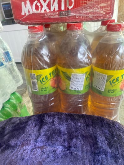 The image shows a close-up view of several plastic bottles filled with iced tea, stacked together. The bottles have yellow labels with green accents, featuring text in what appears to be a Cyrillic or Latin-based script, along with images of peaches. One of the labels prominently displays "ICE TEA" and "shoftoli ta'mi," suggesting a peach flavor. Above the bottles, a red and white packaged product is visible, with Cyrillic text and the words "UZBEKISTAN" printed on it. The bottles are partially wrapped in clear plastic. In the foreground, a dark purple, textured fabric, possibly a towel or blanket, is draped across the lower portion of the image. The background is blurred but suggests an indoor or covered outdoor setting, with hints of other items and surfaces. The overall scene appears to be a retail or storage environment, likely in Kyzyl-Kyya, Kyrgyzstan, given the mention of Uzbekistan on the packaging, indicating potential cross-border trade.