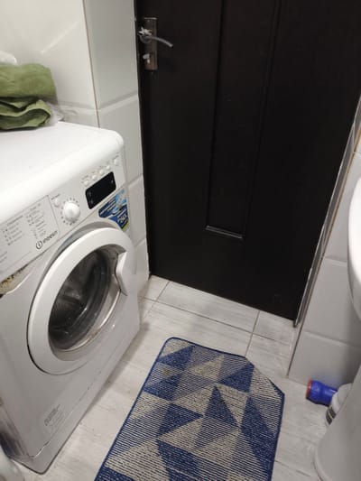 In a dimly lit bathroom in Malinovka, Kyrgyzstan, a white washing machine stands to the left of a dark wooden door. The washing machine has a silver door and a control panel with various buttons and a digital display. A green towel is folded on top of the machine. A blue and white patterned rug lies on the tiled floor in front of the washing machine and a white toilet is partially visible on the right. The walls are tiled in white.