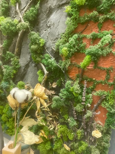 This is a close-up, overhead shot of a decorative arrangement. The primary subjects are various types of green moss, interspersed with dried plants and natural elements. The moss covers a textured, reddish-brown background that resembles brick or clay, and also a dark gray, rough surface. Scattered throughout the moss are several dried, pale yellow flowers, some dried leaves, and a sprig of dried lavender. A single cotton boll is prominently featured near the bottom right. Twigs and branches are also visible, weaving through the moss and plants. The overall impression is that of a natural, somewhat rustic vignette, possibly intended for decoration or a display. There are no people or active events depicted. The lighting appears to be even, suggesting an indoor setting or an overcast day, with no strong shadows. No text is visible in the image.