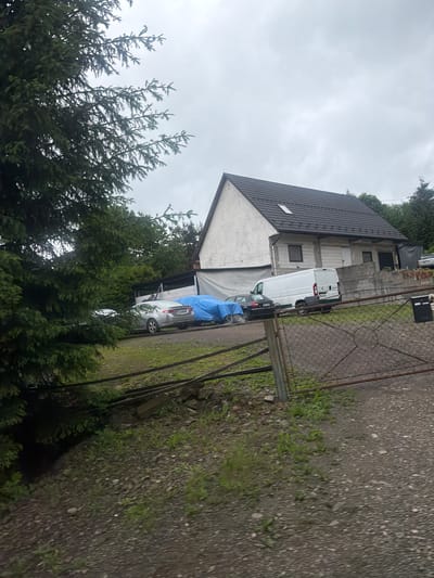 The image depicts a residential property on a cloudy day in Wilkowisko, Poland. A multi-story house with a dark-tiled roof is visible, featuring a small dormer window and a cross symbol above the gable. In front of the house, a gravel driveway leads to a parking area where several vehicles are present. These include a silver sedan partially covered by a blue tarp, a black sedan, a white van, and another silver sedan. A chain-link fence with a rusty metal gate runs across the foreground. Dense evergreen trees are prominent on the left side of the image, partially obscuring the house and vehicles. The ground surrounding the house is a mix of grass and gravel. A black trash bin is located near the gate. The overall scene suggests a quiet, possibly rural or semi-rural, residential setting.