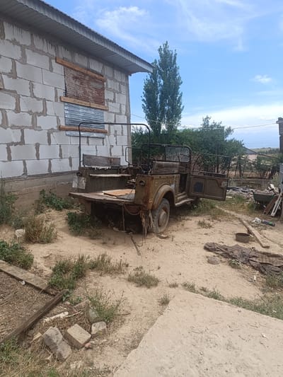 In the city of Shamalgan, Kazakhstan, an outdoor scene depicts a dusty yard with a partially constructed building on the left. The building, made of light-colored cinder blocks, has several boarded-up windows, some covered with corrugated metal and others with rough wood. Adjacent to the building is a rusty, olive-green military-style jeep with its doors open. The jeep appears weathered and worn, with visible rust and a wooden seat in the rear. Various items are scattered around the yard, including lumber, a wheelbarrow, a metal bowl, and some discarded fabric. Sparse vegetation, consisting of dry grass and small green plants, grows in the sandy soil. A chain is visible attached to the rear of the jeep, and a dog is lying underneath it. In the background, trees and distant hills are visible under a blue sky with scattered clouds. The overall impression is one of disuse and neglect.
