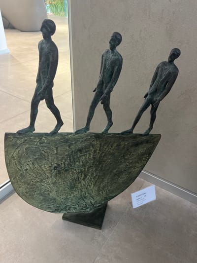 A bronze sculpture by Roberto Barni, titled "Bianco 2" (2002, edition 1/9, 97.5 x 72 x 18 cm, lost-wax technique), is displayed indoors. The sculpture features three elongated, nude male figures with simplified anatomical details, including visible genitalia, cast with a dark greenish patina. They are positioned in a forward-leaning, walking progression atop a textured, semi-circular, boat-like bronze base. The figures face towards the right side of the frame. The leftmost figure is fully reflected in a large, vertical mirror or reflective glass panel positioned adjacent to the sculpture. The sculpture rests on a light-colored, tiled floor. A light brown, subtly textured wall forms the background to the right. An informational label detailing the artwork is placed on the floor to the right of the sculpture. This scene is an indoor art exhibition space, such as a modern gallery or museum hall, located in the city of Galatina, Italy.