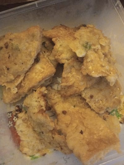The image displays a close-up, high-angle view of numerous irregularly shaped, deep-fried food items contained within a clear, rectangular plastic vessel. The food items exhibit a varied golden-brown to light-yellow coloration with a visibly textured, crispy-appearing surface. Small, dark brown particulates, likely spices or fried herbs, are sparsely distributed across some pieces, alongside occasional faint green fragments embedded within the batter. The items are stacked in a loose pile, occupying the majority of the container's volume. Beneath the food, particularly visible in the bottom left, is a decorative paper liner featuring a repeating pattern of small white and reddish-orange floral motifs on a brown background. The scene is static, without individuals or active interactions, and is consistent with an indoor environment in Kanpur, India.