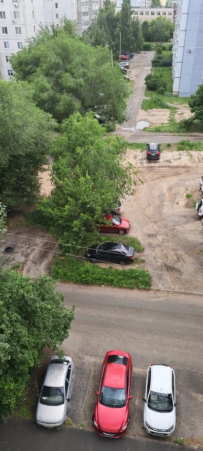 This is a high-angle, outdoor, daylight shot of a parking area next to apartment buildings and trees. The scene appears to be in Syzran, Russia. Several cars are visible, including a silver sedan, a red sedan, a black sedan, a black SUV, and two white cars parked in a line. Some of the cars are parked on a dirt and gravel surface, while others are on asphalt. The trees are lush and green, suggesting it's a warm season. There's a paved path leading to the parking area, with a puddle of water on it, indicating recent rain. The buildings in the background are typical multi-story apartment blocks. No people are visible. The overall impression is of a residential area during the day.