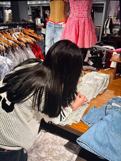 A person with long, dark hair stands in a clothing store. Racks of clothing are visible on the left side of the frame, with hangers and various garments displayed. On the right, a wooden table showcases folded clothes and denim items. The person is wearing a white, patterned sweater with black accents, their hands occupied with examining a piece of clothing on the table.