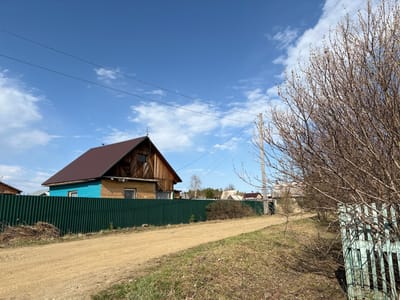 The image shows a rural scene, seemingly in the Ust'-Ilimsk area of Russia. A dirt road runs parallel to a green corrugated metal fence. Beyond the fence are houses, one with brown wooden siding and a reddish-brown roof, and another with blue siding. Above the houses, a blue sky with scattered clouds is visible. In the foreground, a leafless tree with budding branches is present. A weathered, light blue wooden fence can be seen on the far left of the image. Power lines stretch across the sky.