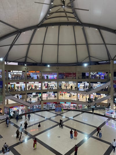 This image depicts the interior of a large shopping mall in Kalyan-Dombivli, India. The mall features a multi-story structure with various shops and brands visible on different levels, including Burger King, Barbeque Nation, Easybuy, Max, Croma, and Smart Bazaar. Escalators are visible, connecting the different floors. People are seen walking and shopping on the patterned tile floor. The mall has a distinctive domed ceiling with visible support beams and lighting fixtures.
