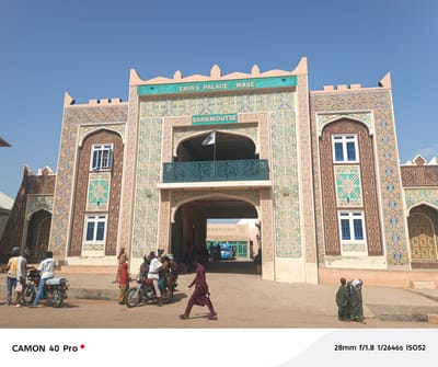 The image shows the Emir's Palace Wase in Wase, Nigeria. The palace is a large, intricately decorated structure with arches, windows, and a sign that reads "EMIR'S PALACE WASE" and "SARKIN DUTSE" above the main entrance. People are present in the foreground, some standing near motorcycles and others walking towards the palace entrance. A building is visible through the palace's archway. The sky is clear and blue. The image capture settings are displayed at the bottom of the picture.