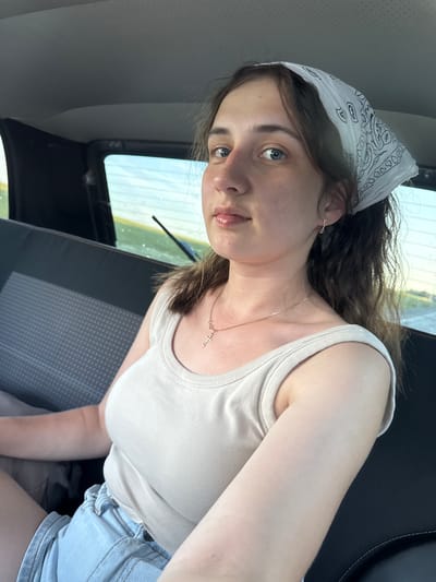 A young woman with blue eyes, brown hair, and a white bandana on her head is seated in the back seat of a vehicle. She is wearing a light-colored tank top and denim shorts, and a gold cross necklace. The woman is looking at the camera, and her lips are slightly parted, revealing her tongue. The interior of the car is visible, including the dark gray upholstered seat and the window, which shows a blurred background of a body of water and a sky. The scene appears to be in Verkh-Sosnova, Russia.