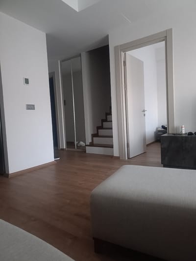The image depicts an interior view of a modern residence in Urla, Turkey. The scene includes a wooden floor leading to a hallway with a set of stairs. To the left, a white wall features a thermostat and light switches. A mirrored closet door is visible in the hallway, reflecting a pair of white sneakers. An open doorway on the right reveals a white door and a dark cabinet with various items on top, including a bottle of spray and sunglasses. In the foreground, a portion of a light-colored ottoman is visible. The overall impression is of a clean and contemporary living space.
